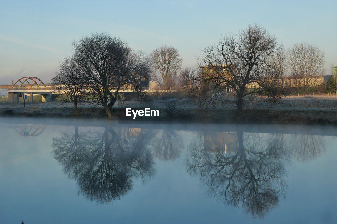 reflection, tree, water, morning, winter, sky, plant, lake, nature, bare tree, tranquility, no people, beauty in nature, tranquil scene, scenics - nature, architecture, landscape, built structure, dawn, waterfront, snow, outdoors, day, reservoir, blue, clear sky, mist