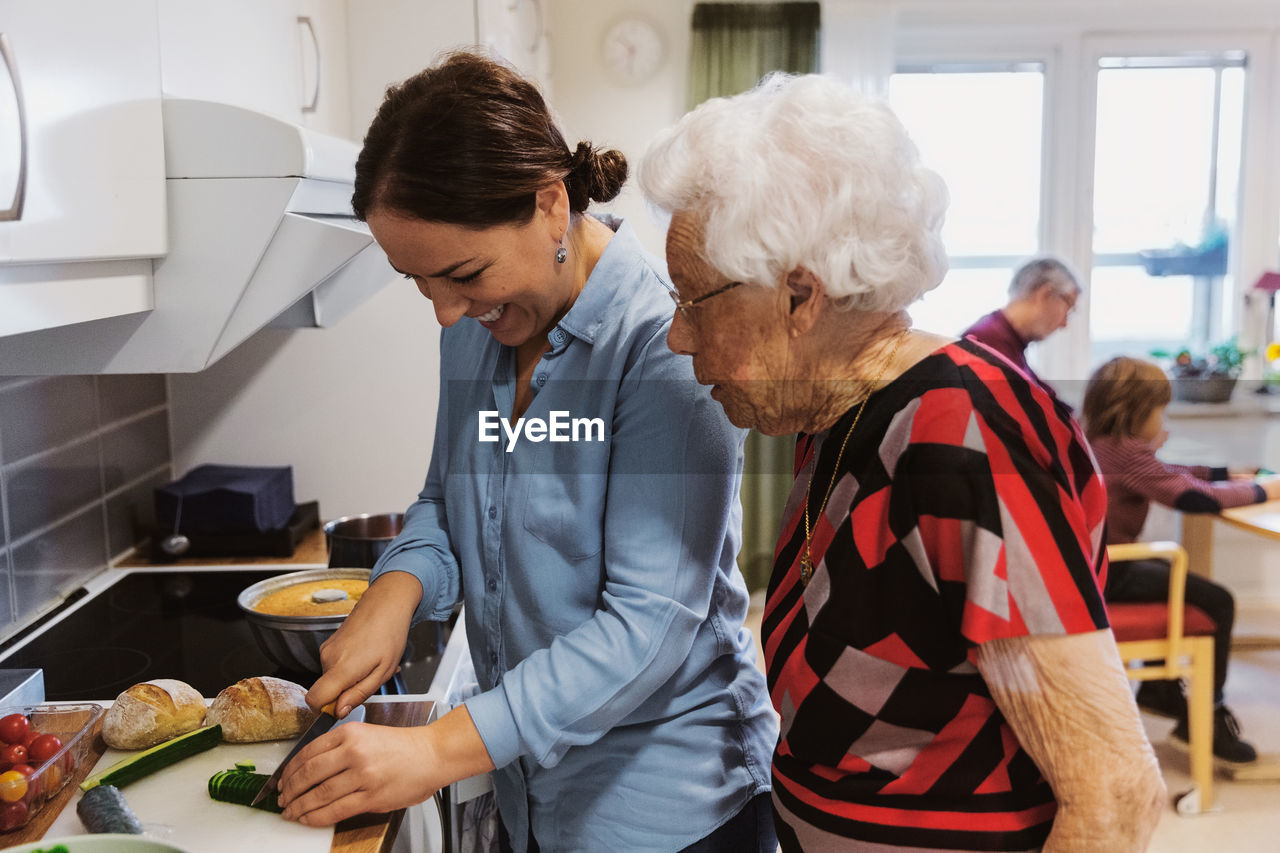 Side view of senior woman looking at daughter cutting zucchini in kitchen