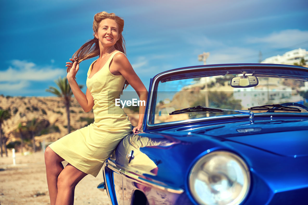 Portrait of smiling young woman sitting on car against blue sky