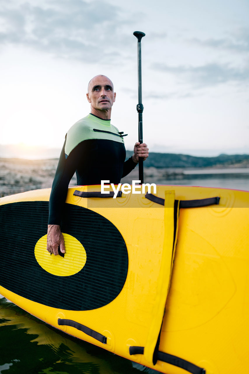 Side view of male surfer in wetsuit standing with yellow sup board and paddle in sea water