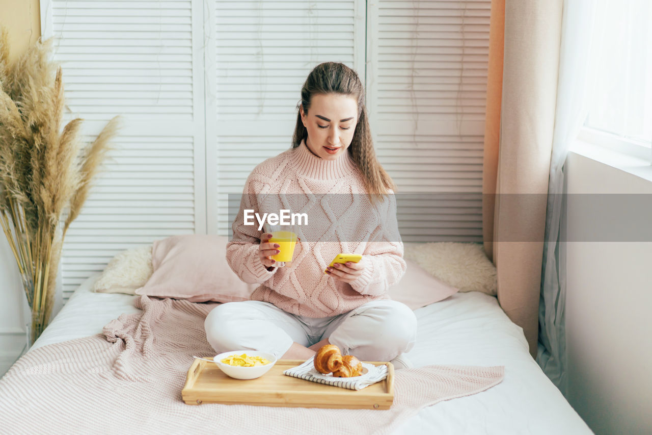 Smiling woman using mobile phone while sitting on bed by breakfast at home