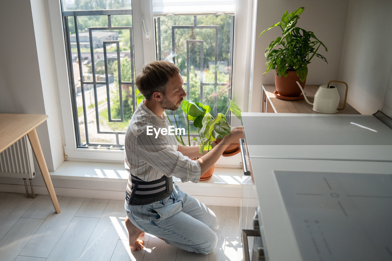 Young man wearing back support belt taking care of houseplants at home