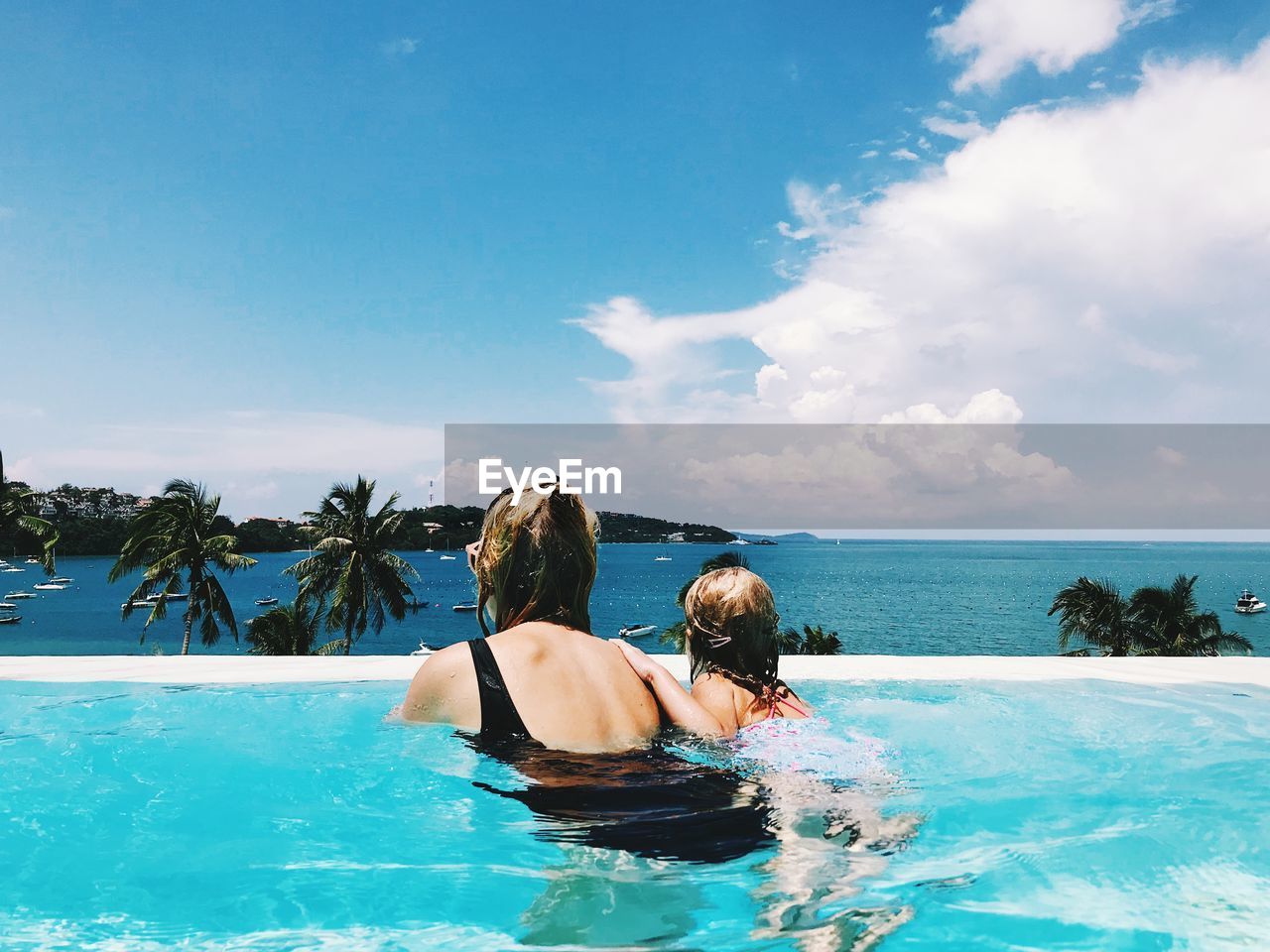 Rear view of woman with daughter swimming in pool by sea against cloudy sky