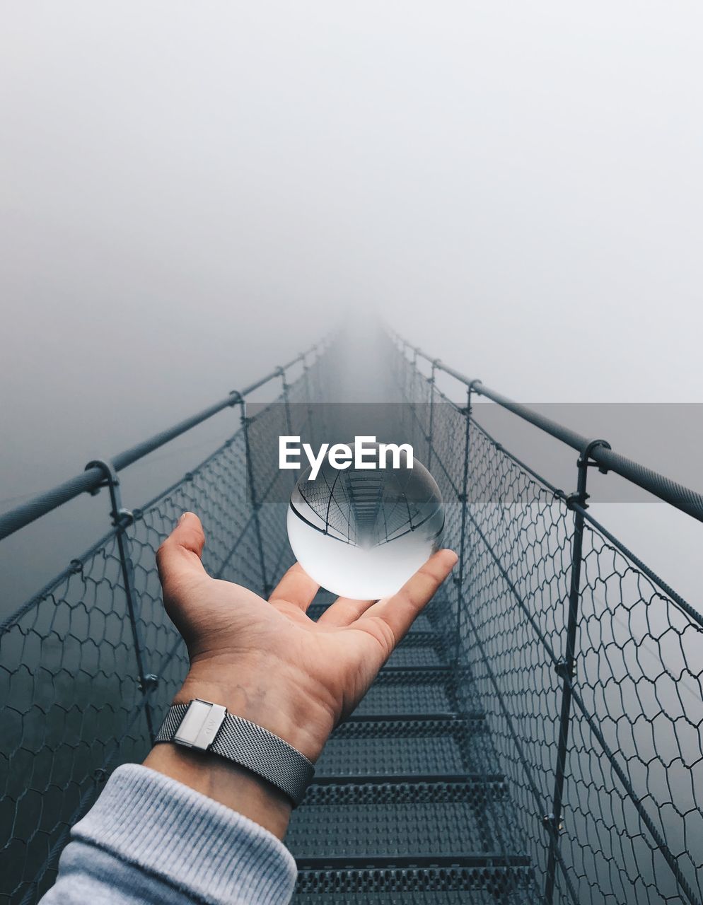 Cropped hand of man holding crystal ball on footbridge against fog