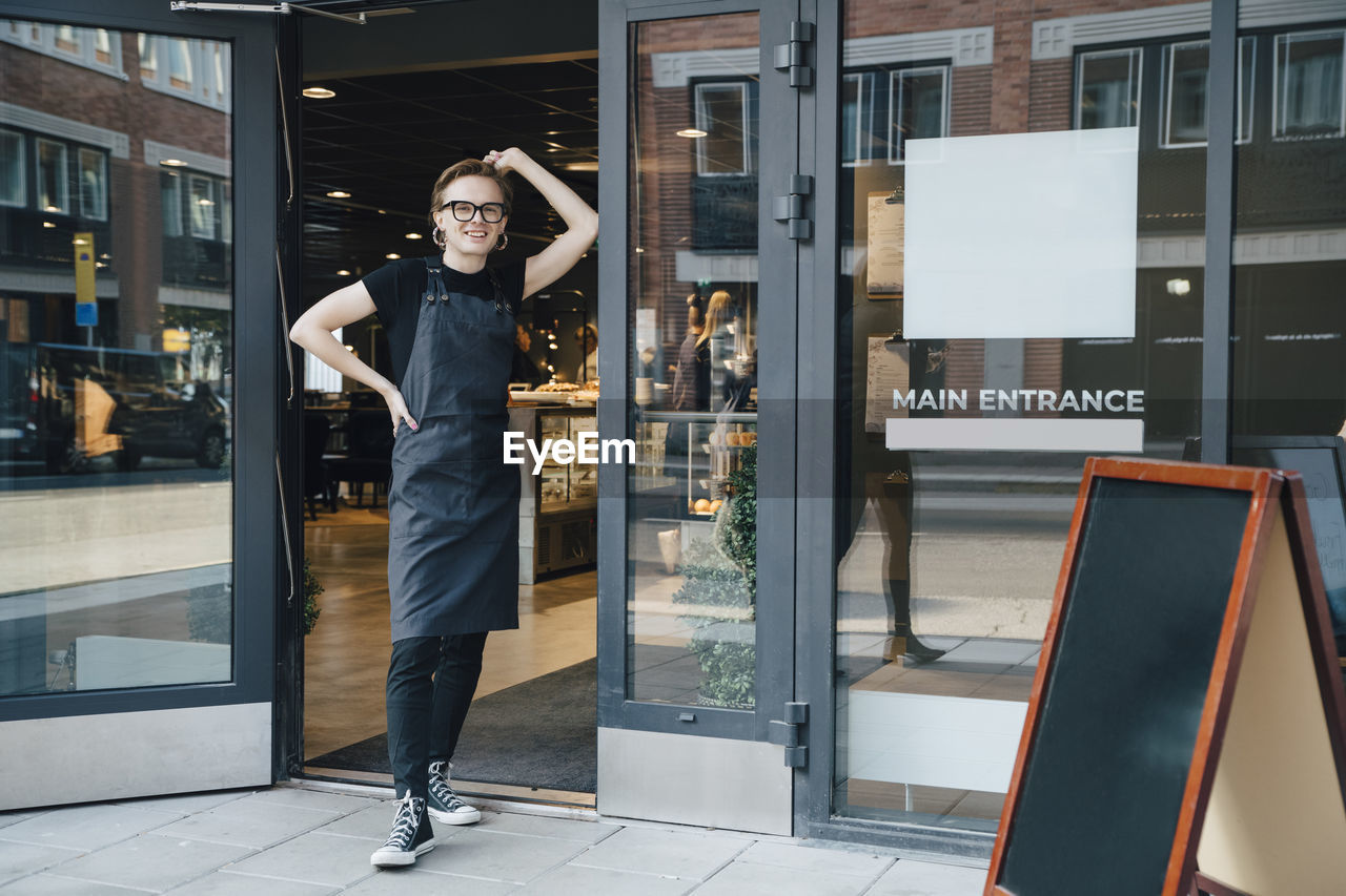 Full length portrait of smiling young owner standing at cafe entrance