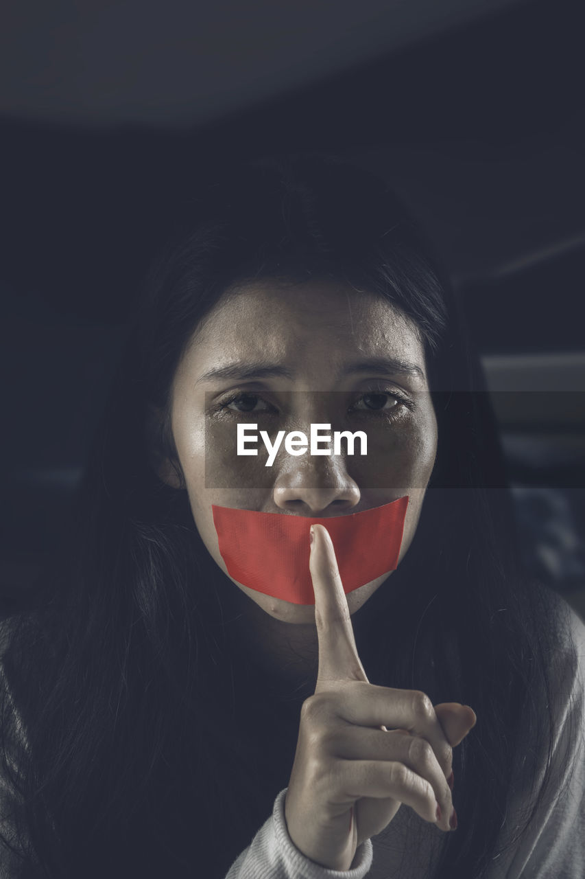 Close-up portrait of young woman gesturing over tape on mouth in darkroom