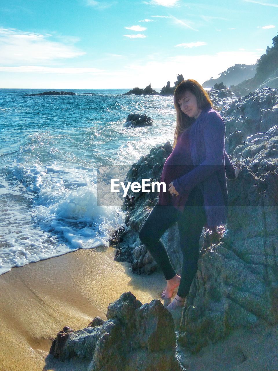 Full length of pregnant young woman standing by rocks on shore at beach