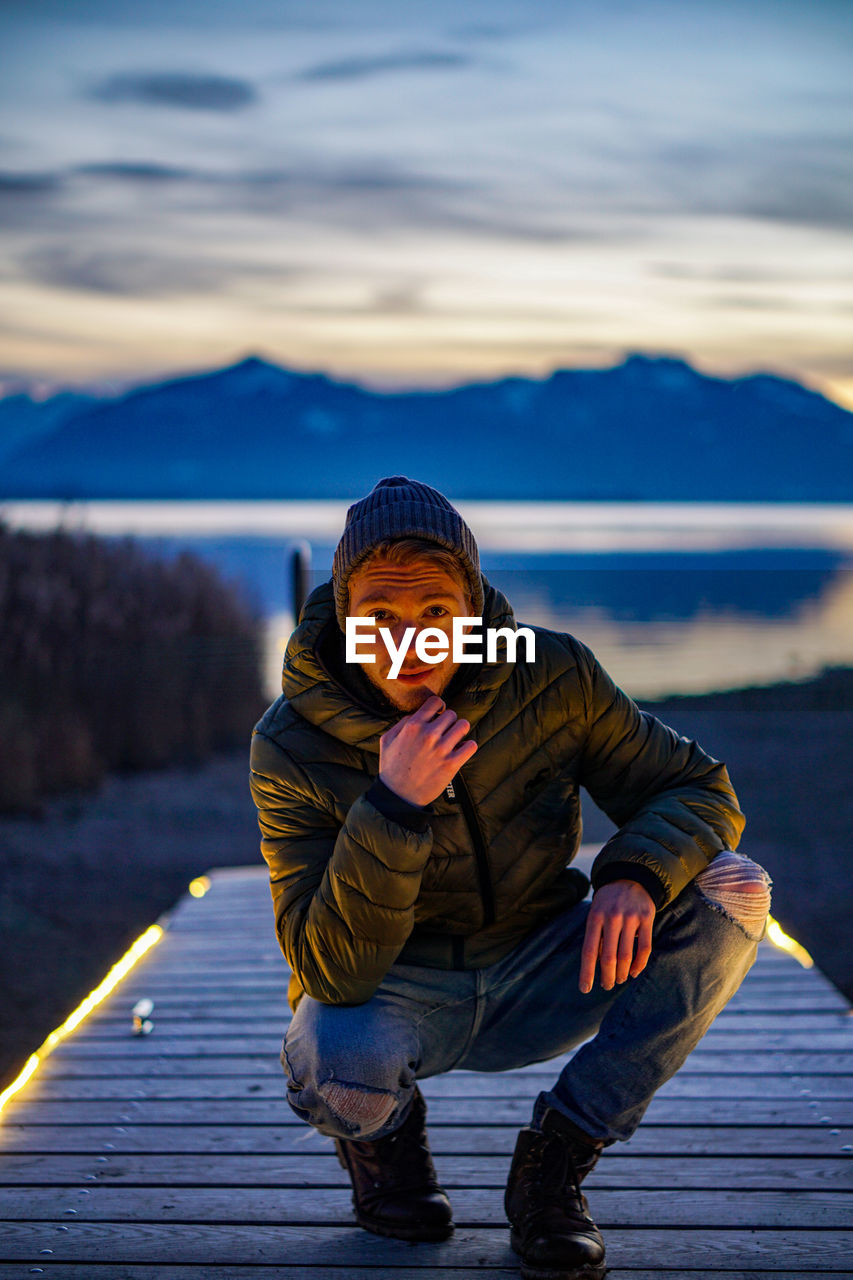 Portrait of young man crouching on pier against lake