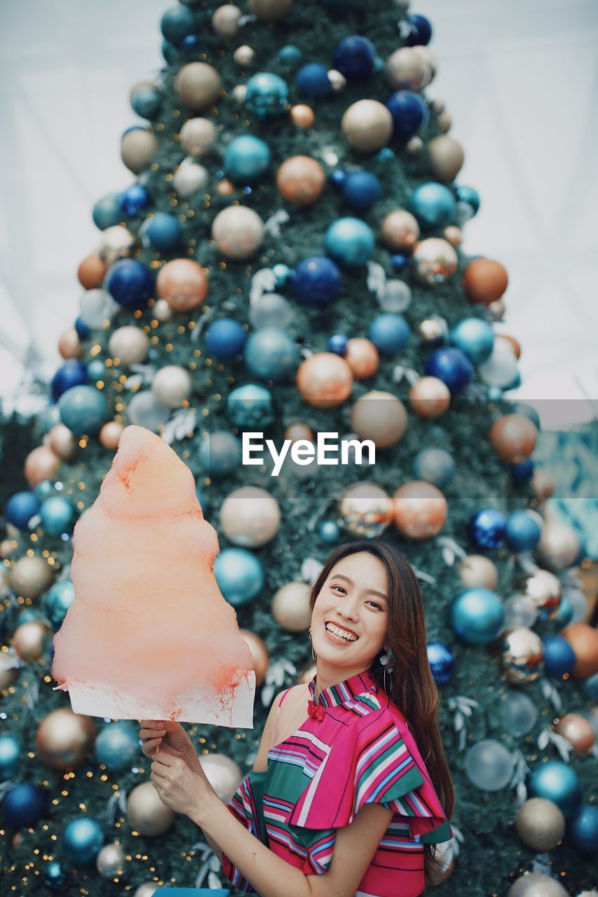 Portrait of smiling young woman standing by christmas tree holding cotton candy outdoors