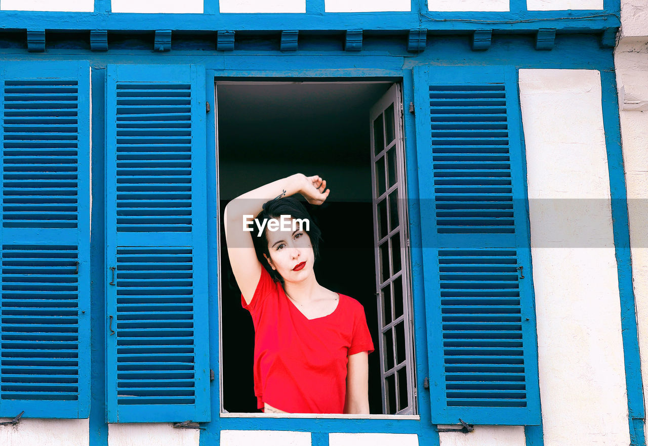 PORTRAIT OF YOUNG WOMAN STANDING BY WINDOW OF BUILDING