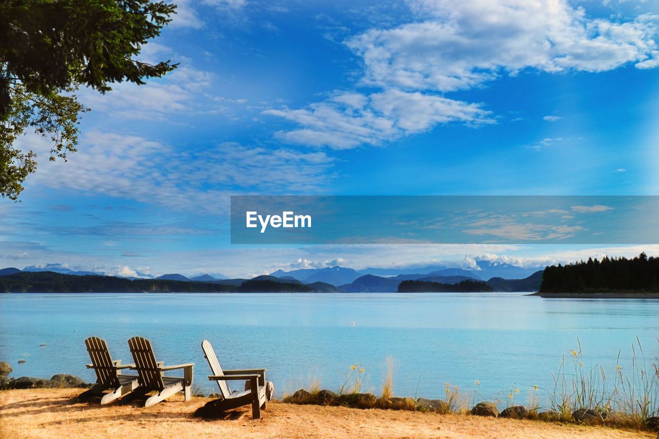 Empty chairs by lake against sky