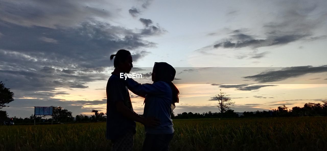 sky, two people, sunset, cloud, adult, silhouette, nature, men, togetherness, emotion, horizon, love, field, positive emotion, child, plant, women, sunlight, bonding, person, childhood, landscape, family, land, standing, evening, outdoors, dusk, female, embracing, parent, grass, environment, three quarter length, lifestyles, back lit, one parent