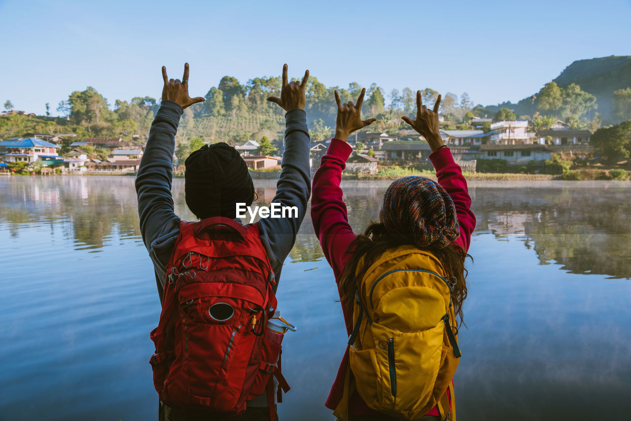 Rear view of people gesturing while standing by lake against sky