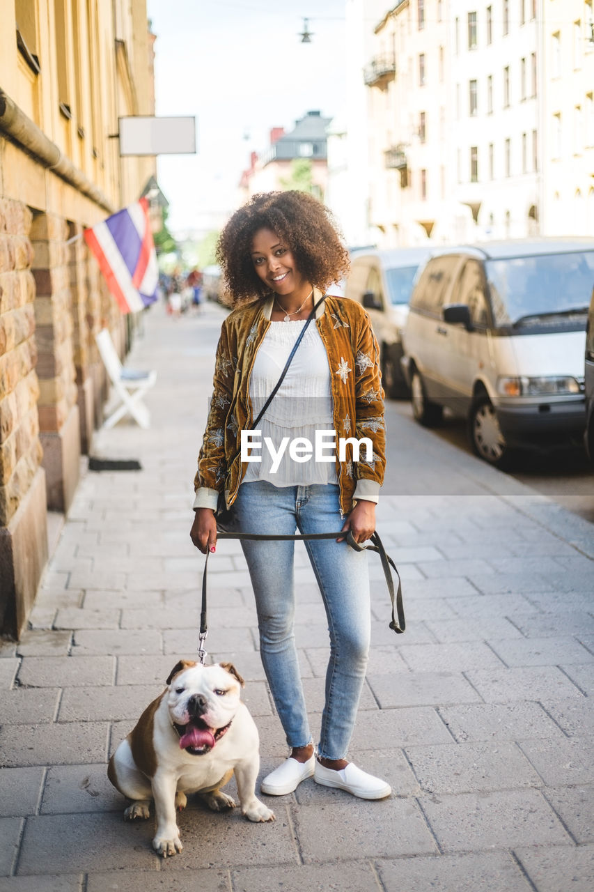 Portrait of smiling mid adult woman standing with dog on sidewalk in city