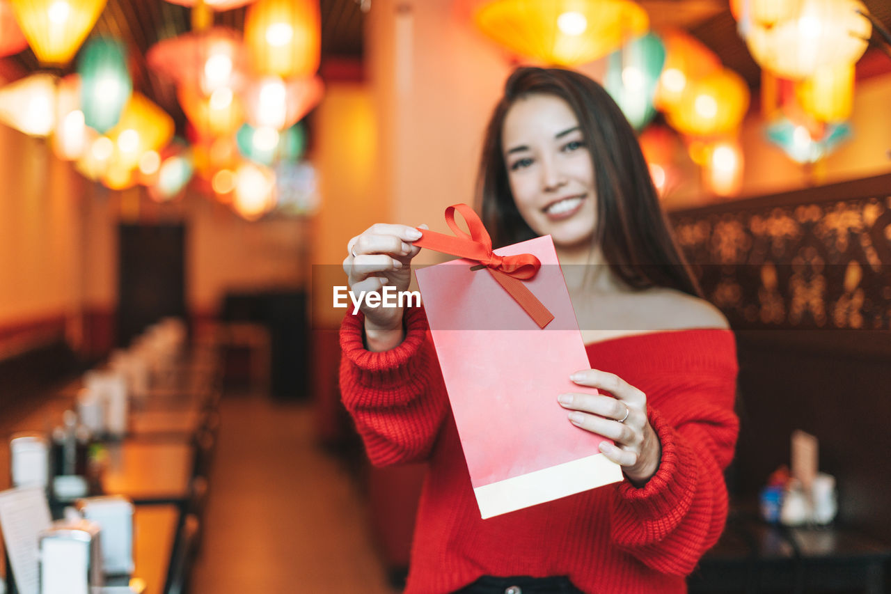 Beautiful young asian woman in red clothes with red gift bag in restaurant celebrating 
