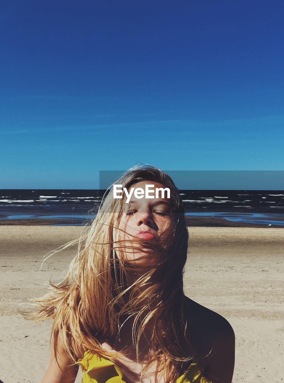 Young woman with eyes closed sticking out tongue while standing at beach against blue sky