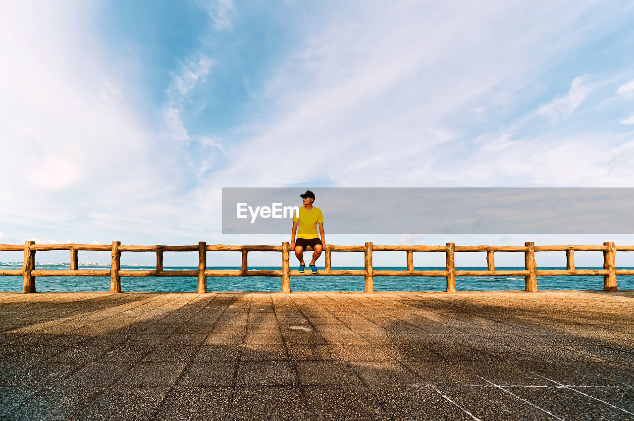 Man sitting on railing at promenade against cloudy sky