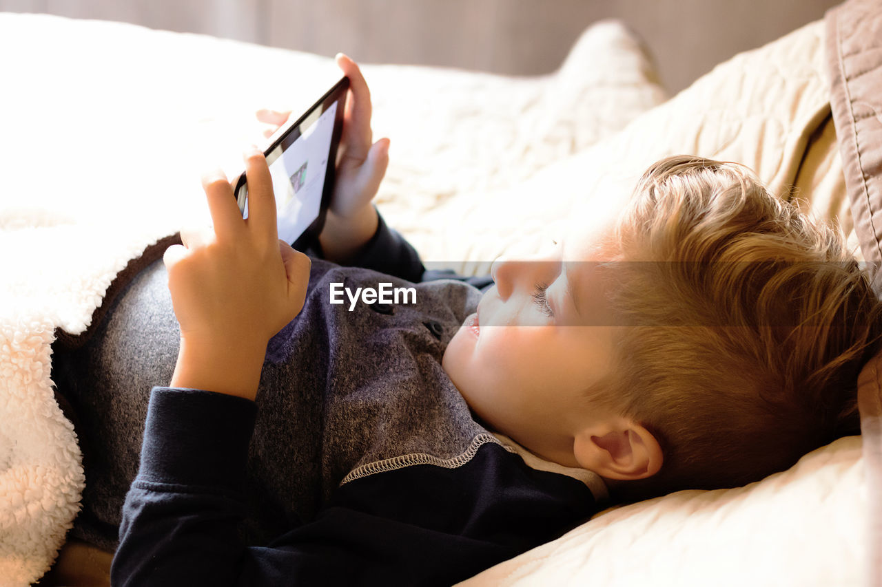 Small boy using touchpad while lying down in bedroom.