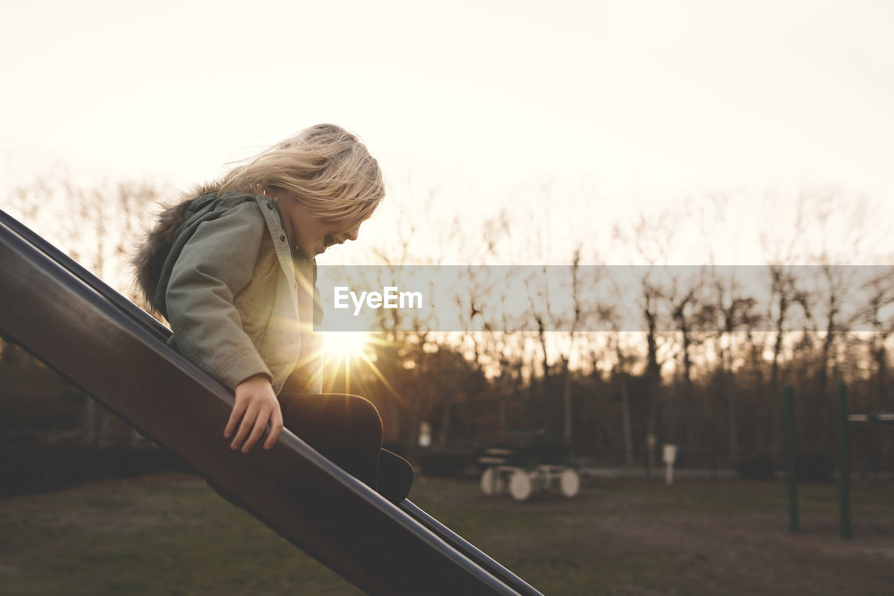 Side view of cheerful girl playing on slide at playground during sunset