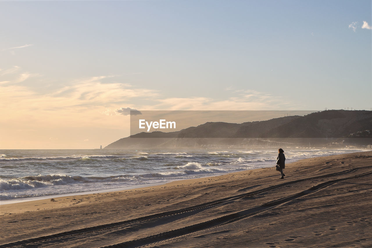 Woman standing at beach against sky during sunset
