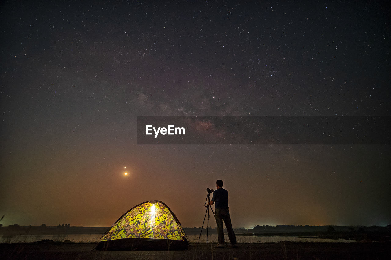 Rear view of silhouette man with camera standing against sky at night