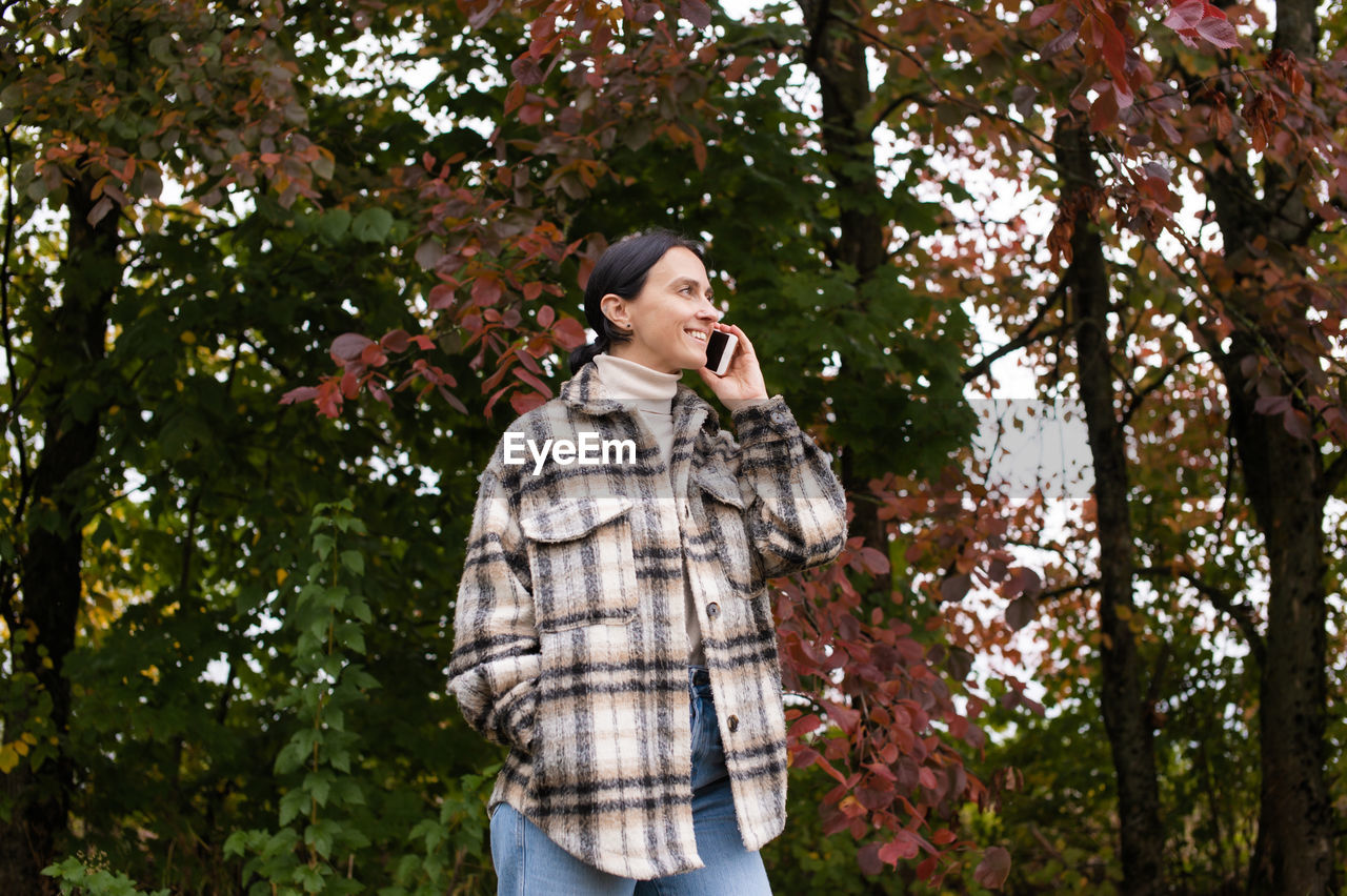 A girl in warm clothes is talking on the phone against the background of the forest and laughing