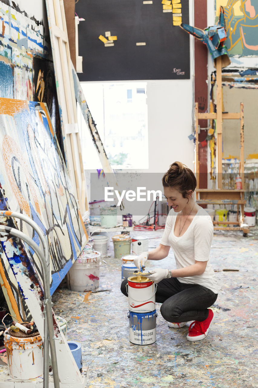 Happy artist crouching while dipping brush in paint can at art studio