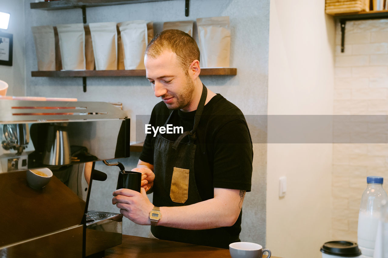 Smiling male barista in apron making coffee and whipping milk in coffee machine in cafe