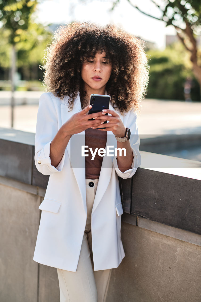 Hispanic female entrepreneur in white suit with curly hair browsing on smartphone while standing near fence on sunny day on city street