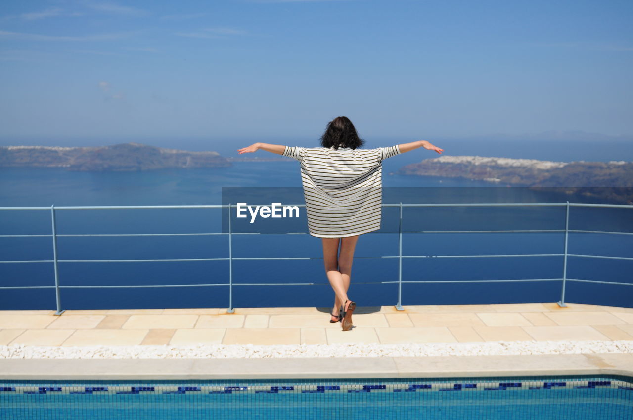 Rear view of woman standing with arms outstretched by railing at poolside against sea