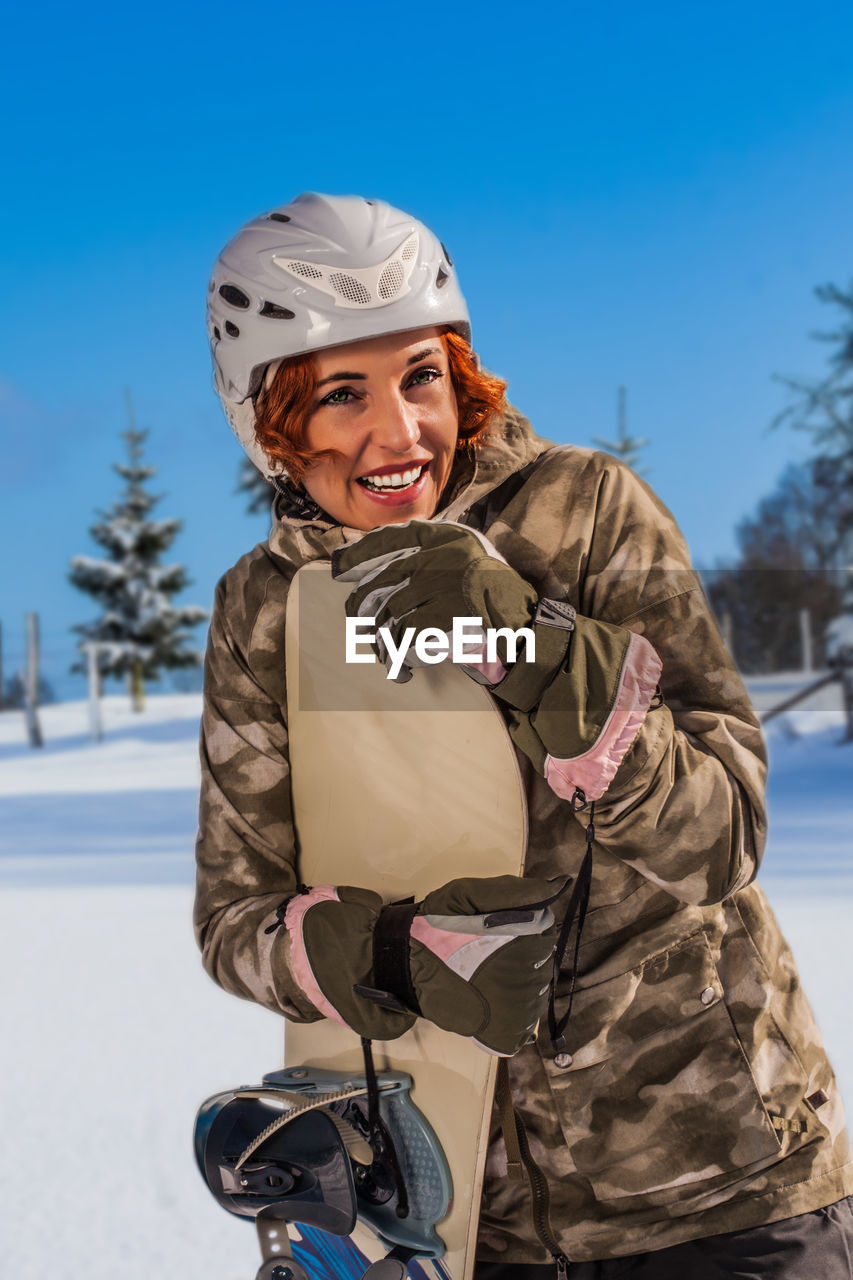 Smiling woman with snowboard looking away while standing on snow covered field against blue sky