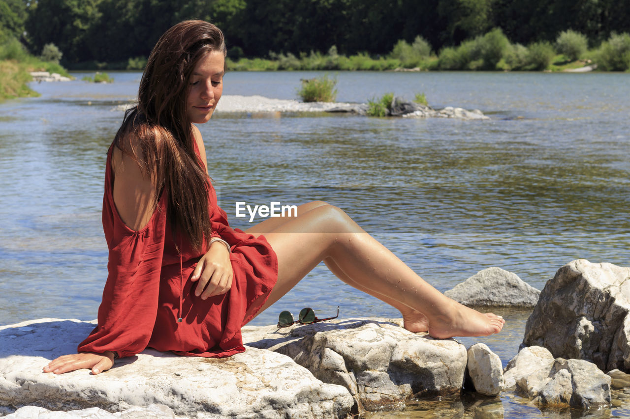BEAUTIFUL YOUNG WOMAN SITTING ON ROCK BY LAKE