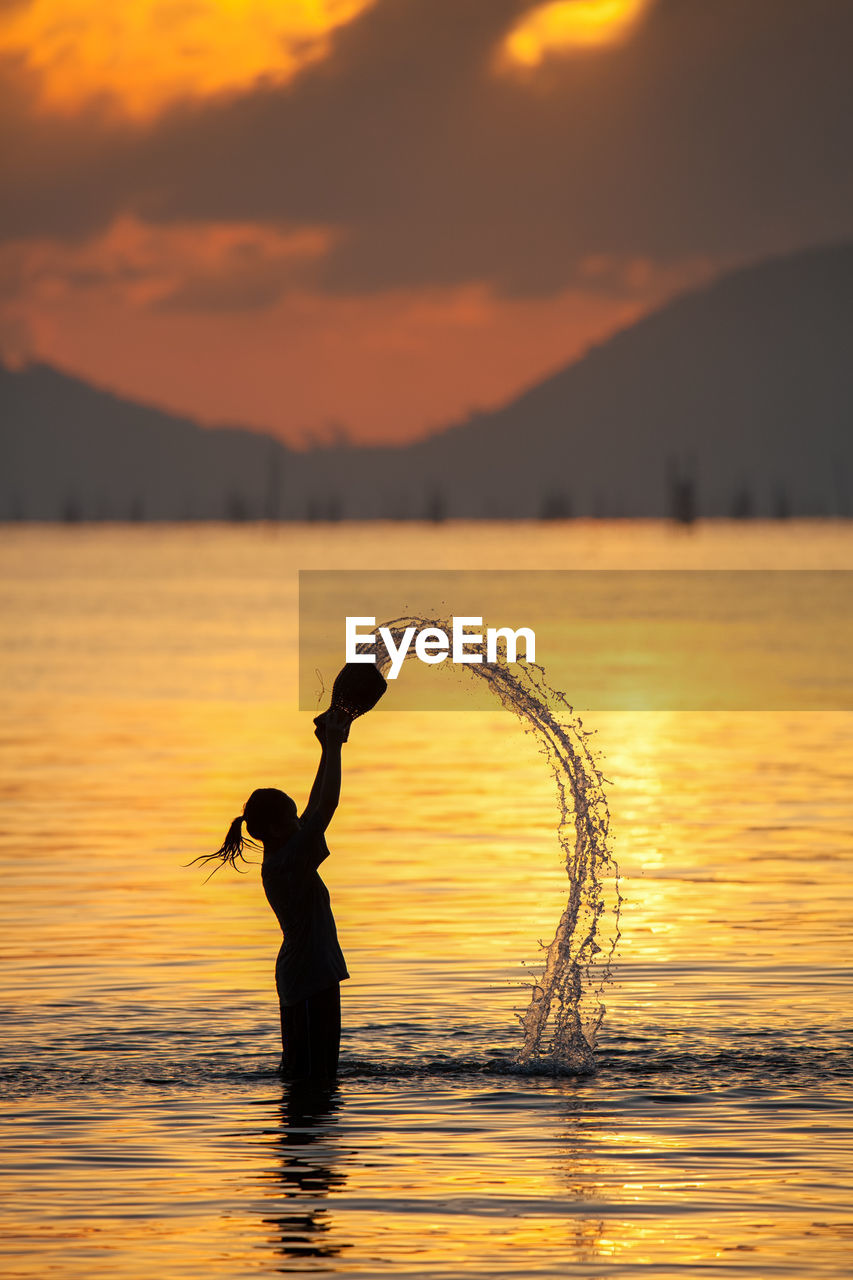 Side view of silhouette woman throwing water in lake against mountains during sunset