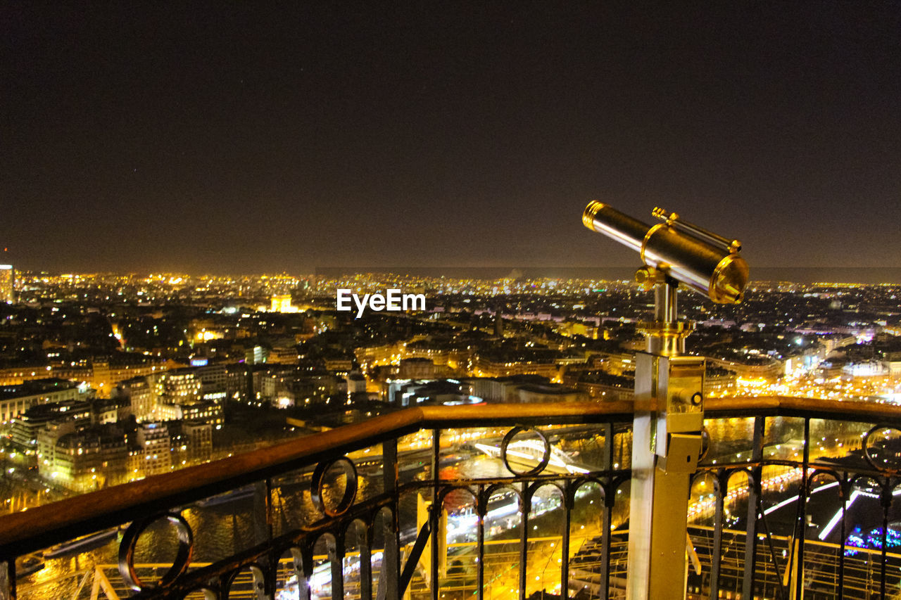 Telescope in balcony against illuminated buildings in city at dusk