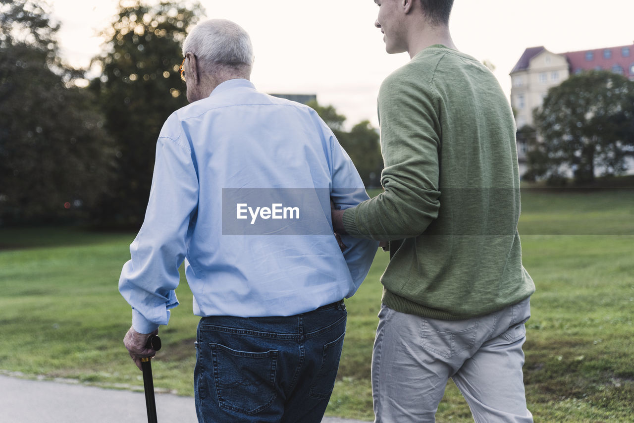 Back view of young man assisting his grandfather walking in a park