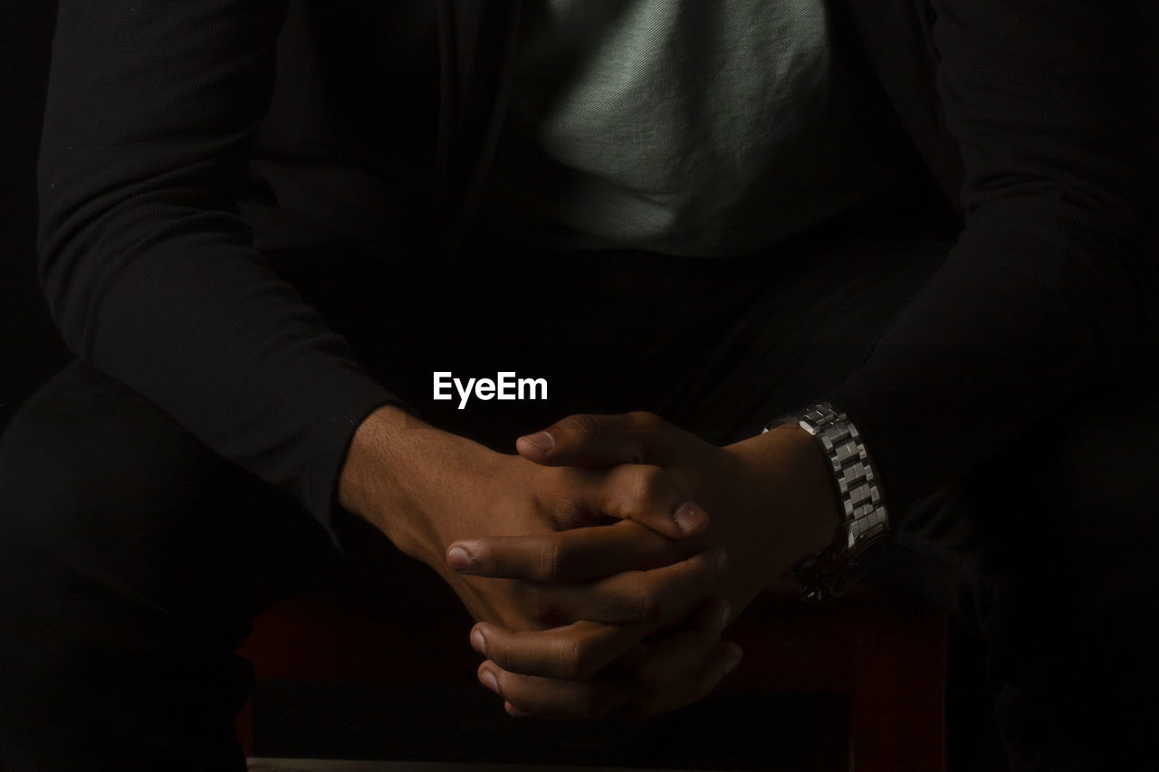 Midsection of man sitting with hands clasped against black background