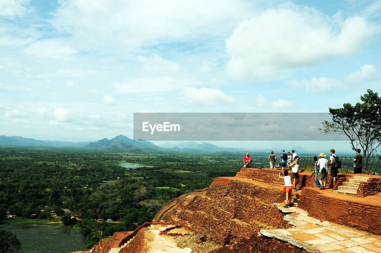 Tourists on lion rock looking at view of mountains against cloudy sky