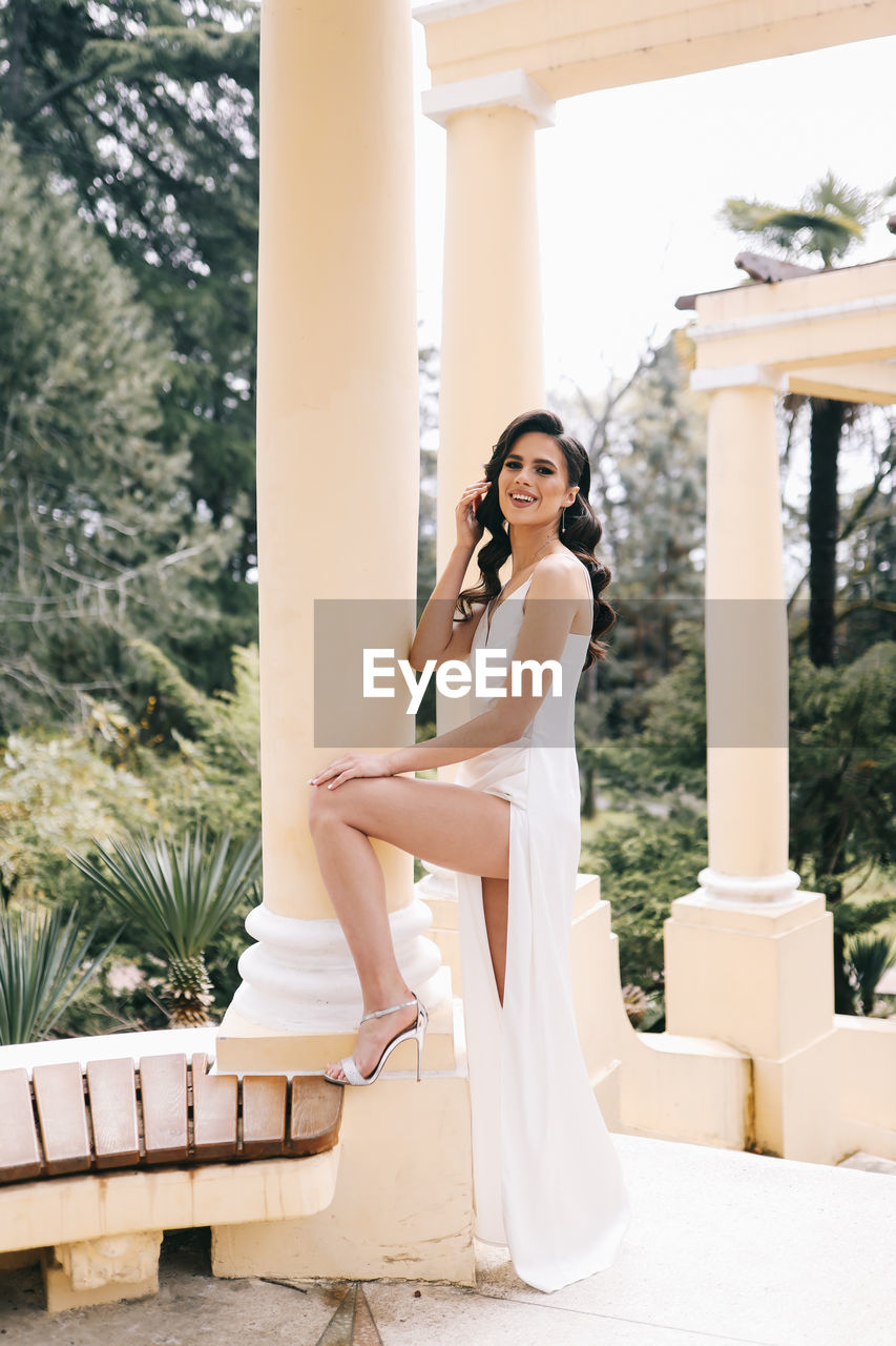 A beautiful brunette lady in an elegant wedding dress poses among the columns in the old city park