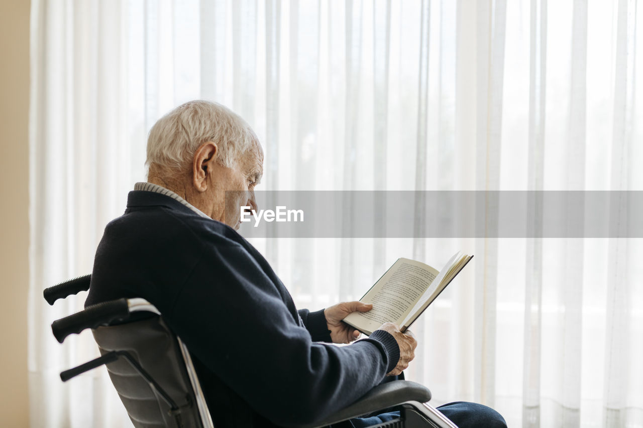 Senior man sitting in wheelchair reading a book near the window