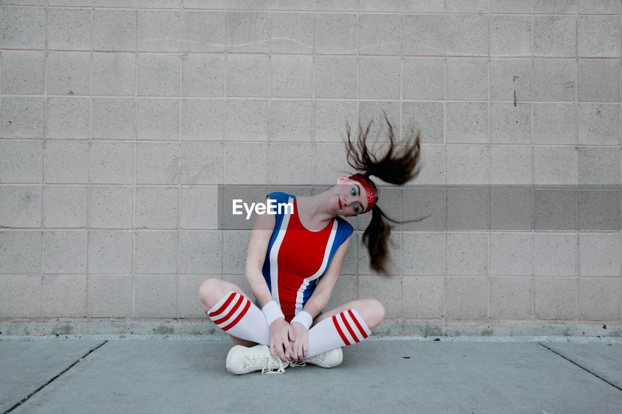 Woman in sports costume tossing hair while sitting on sidewalk against wall