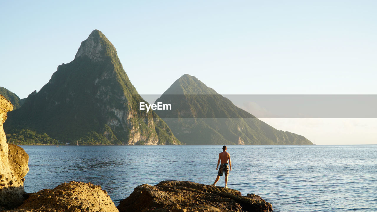 Rear view of shirtless man standing on rock by sea