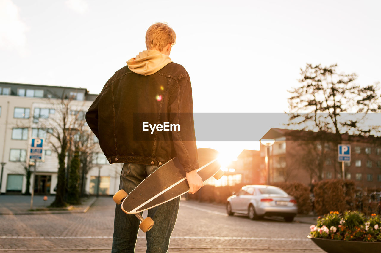 Rear view of young man holding skateboard while standing on sidewalk in city