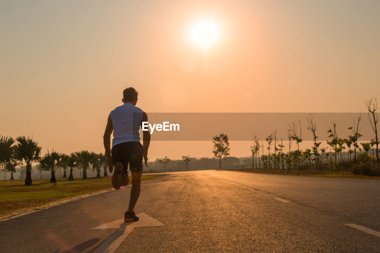 REAR VIEW OF MAN RUNNING ON ROAD AGAINST SKY AT SUNSET