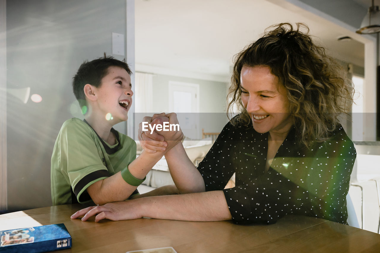 Playful mother arm wrestling with son on table at home