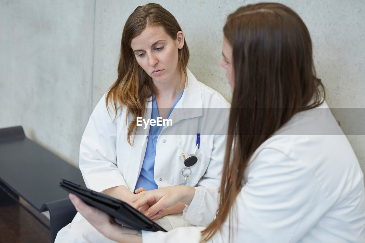 Confident young female doctor discussing with coworker over digital tablet while sitting against wall at hospital