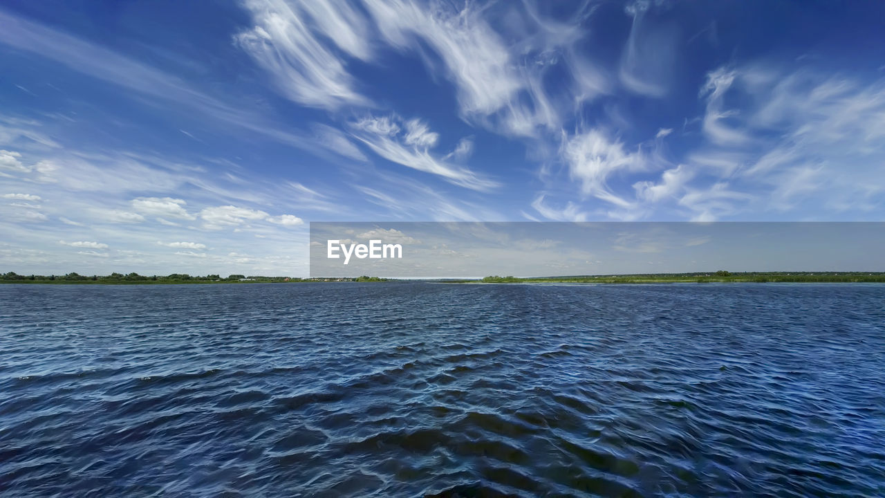 Blue sky with white cirrus clouds, turquoise water, green coastline on the horizon. 