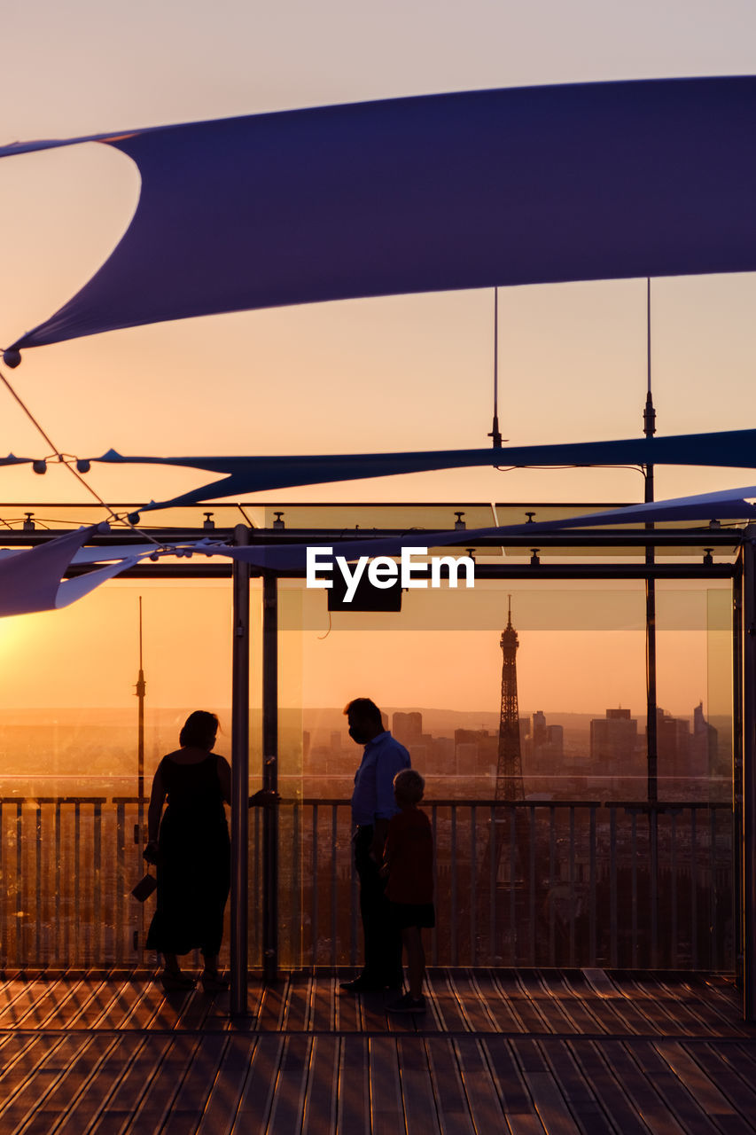 REAR VIEW OF MAN STANDING BY RAILING AGAINST SKY DURING SUNSET