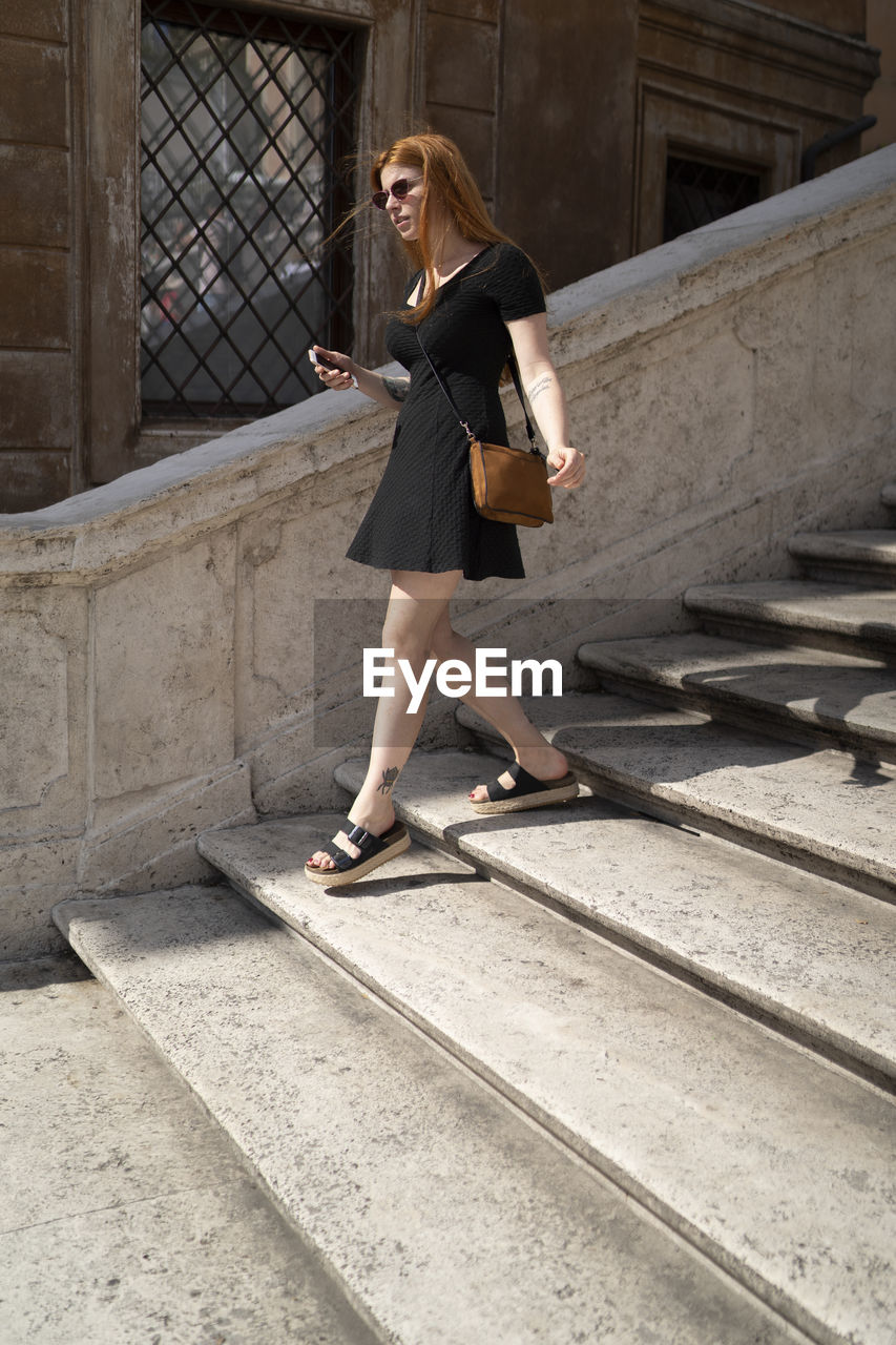 Red ginger woman in black dress descending stairs holding a smartphone