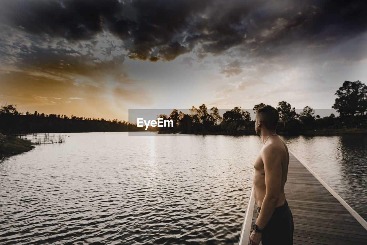 Side view of shirtless man standing by lake on jetty during sunset