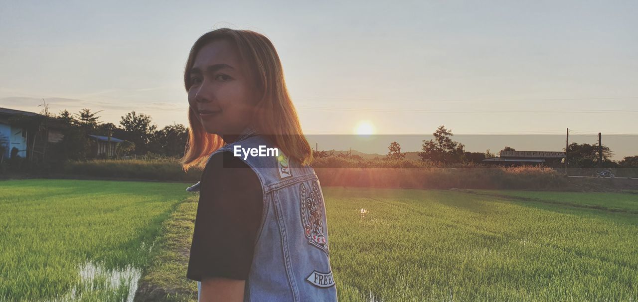 one person, sky, nature, field, sunset, women, plant, adult, sunlight, young adult, grass, standing, landscape, architecture, land, casual clothing, lens flare, hairstyle, lifestyles, rural scene, long hair, environment, built structure, outdoors, waist up, leisure activity, back lit, portrait, flower, agriculture, female, beauty in nature, rural area, child, building exterior, growth, person, sunbeam, looking, tranquility, tree, day, copy space, looking away, sun, childhood, dusk, farm, contemplation